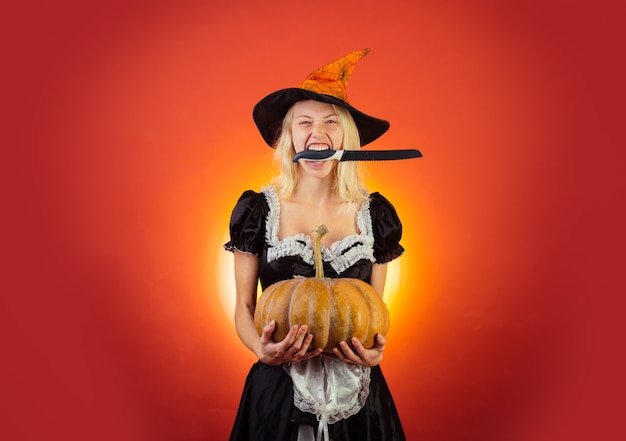 Close-up portrait of gorgeous happy blonde fairy enchantress, covering her eyes with the cap, hot figure, body. Beautiful young surprised woman in witches hat and costume holding pumpkin.