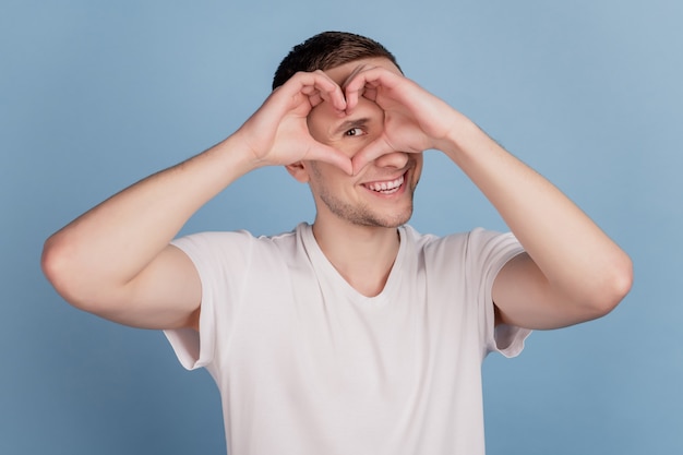 Ritratto ravvicinato di un ragazzo sorriso felice che mostra il segno del cuore il giorno di san valentino guarda l'occhio isolato su sfondo blu