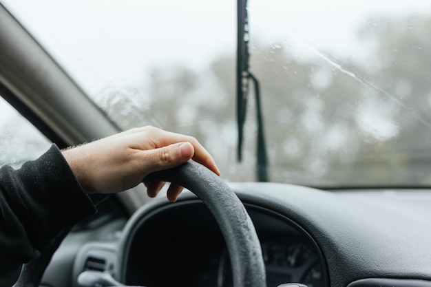 霧と雨の冬の日の旅行中に車のハンドルを運転している認識できない男の手の肖像画をクローズアップ