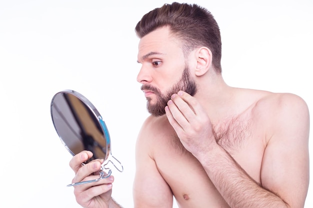 Close up portrait of a handsome bearded man with towel looking at his reflection in the mirror in bathroom isolated on white background