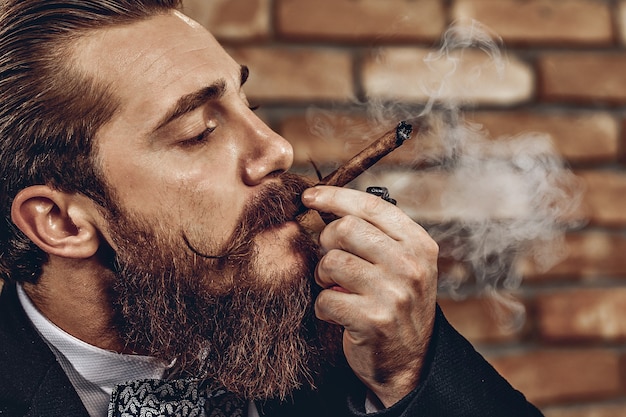 Close up portrait of handsome brutal man with a mustache and beard smoking a brown cigar against a brick wall background. Smoke concept