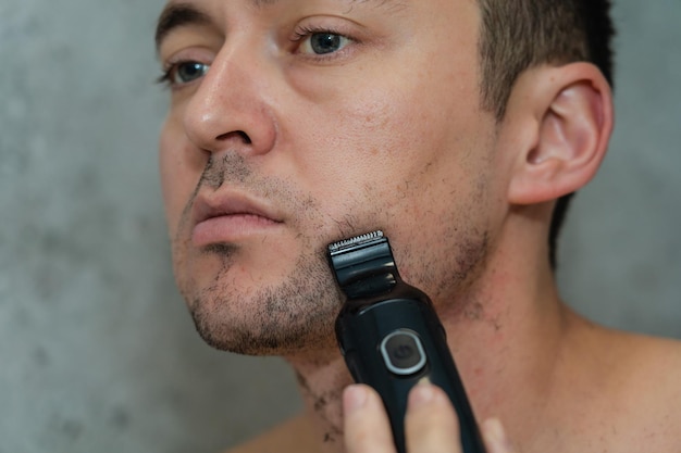 Close up portrait on handsome man shaving his beard by electric trimmer Morning hygiene