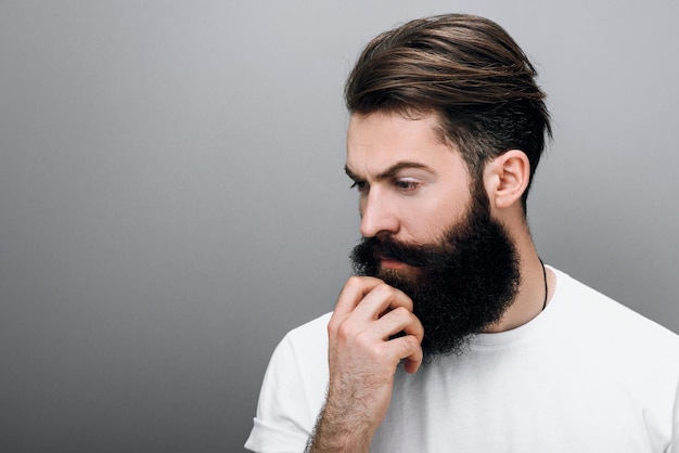 Close up portrait of handsome masculine young bearded man is keeping hand on beard and looking down on a gray studio background with copy space Portrait of young European hipster with trendy beard