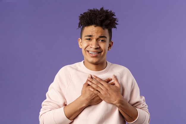 Close-up portrait of handsome silly hispanic guy with dreads, place hands on heart and sighing as contemplate something adorable speaking from all his soul, thanking for praises, purple background