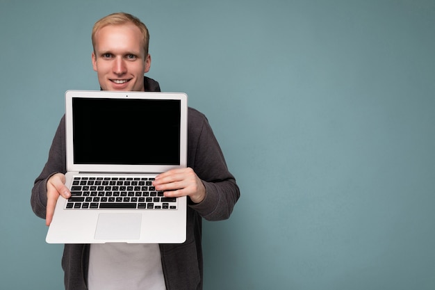 Close-up portrait of handsome smiling blonde man holding computer laptop with empty monitor screen with mock up and copy space looking camera isolated over blue background