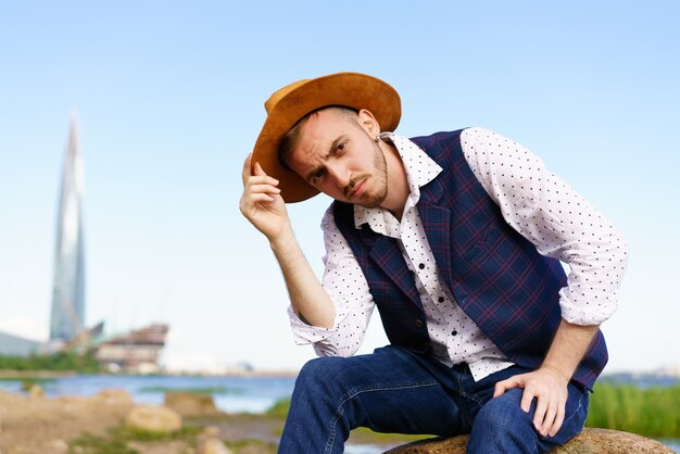 Close up portrait of handsome stylish casual man traveler in hat sitting at sea coastline and enjoying peaceful nature