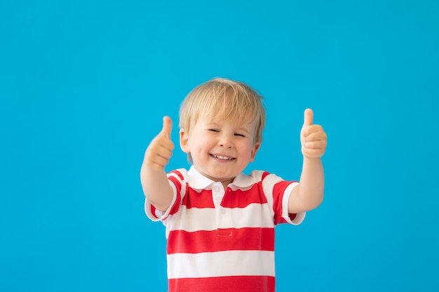 Close up portrait of happy child wearing striped shirt showing thumbs up against blue wall.