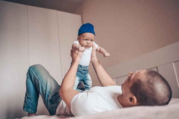 Close up portrait of happy young dad father holding his baby in blue jeans and white t-shirt and cap. Young happy family, dad playing with cute emotional little newborn child son in the bedroom.