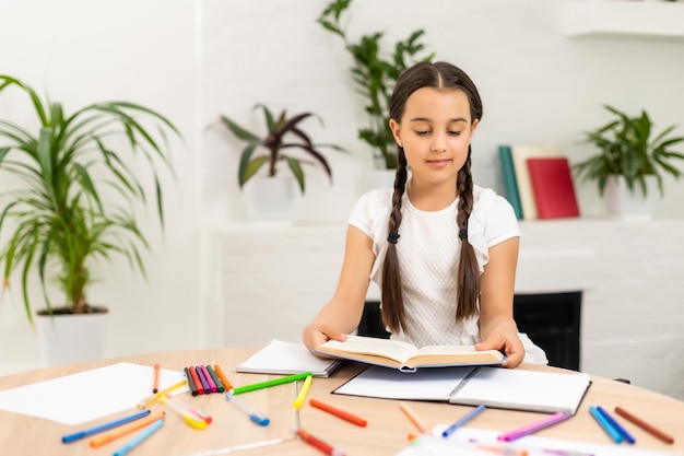 彼女のクローズアップの肖像画彼女は素敵な魅力的なかわいい小さな陽気な長髪の女の子座っているテーブルデスクトップライティング記事タスク宿題屋内の明るい白い教室で。