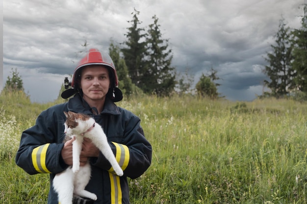 Крупным планом портрет героического пожарного в защитном костюме и красном шлеме держит на руках спасенную кошку. Пожарный в операции по тушению пожара. Фото высокого качества