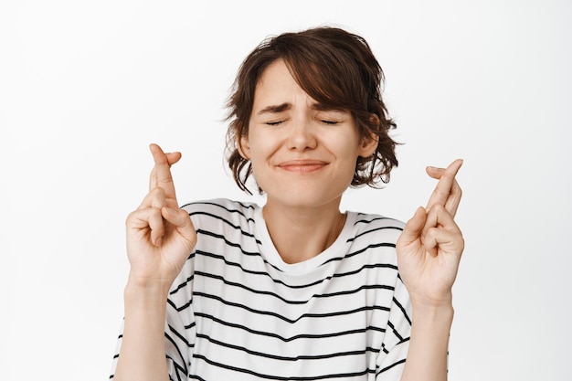 Close up portrait of hopeful nervous girl close eyes, smiling with fingers crossed, making wish, praying, standing over white background.