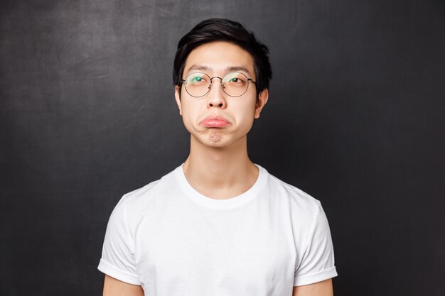 Close-up portrait of impressed asian young man in white t-shirt and glasses, look upper left corner with approval say not bad, squinting and nod acceptingly, standing black wall