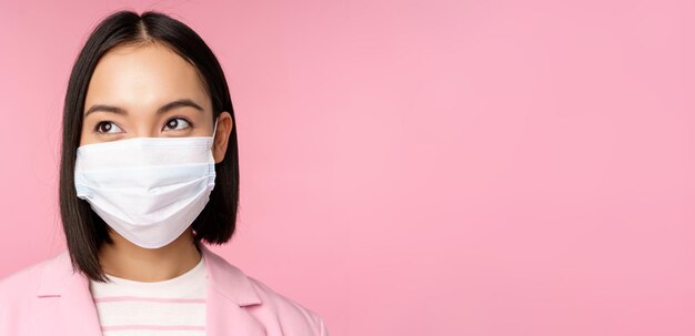 Photo close up portrait of japanese corporate woman in medical face mask from covid-19 looking left at logo sale promo standing over pink background                               person