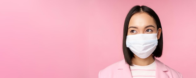 Крупный план портрета японской корпоративной женщины в медицинской маске от covid19, смотрящей влево на промо-акцию по продаже логотипа, стоящую на розовом фоне