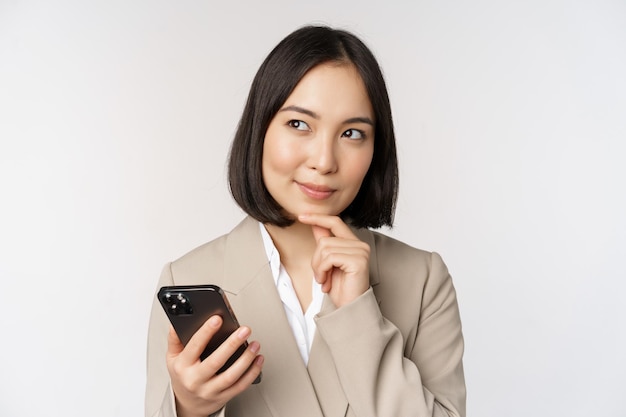 Primo piano ritratto di donna coreana donna aziendale in tuta utilizzando il telefono cellulare e sorridente tenendo lo smartphone in piedi su sfondo bianco