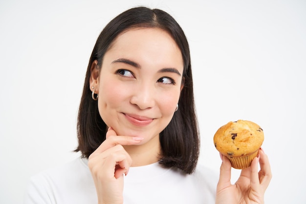 1 つのカップケーキを考え、白いスタジオの背景に笑顔を持つ韓国人女性の肖像画を間近します。