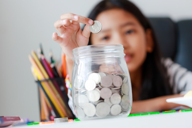 Close up portrait little Asian girl putting money coin into the glass jar piggy bank select focus shallow depth of field