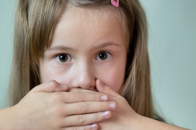 Close-up portrait of little child girl with long hair covering her mouth with hands.