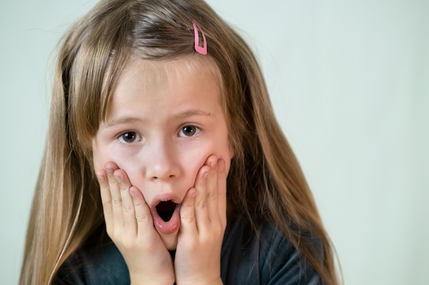 Close-up portrait of little child girl with long hair covering her mouth with hands.