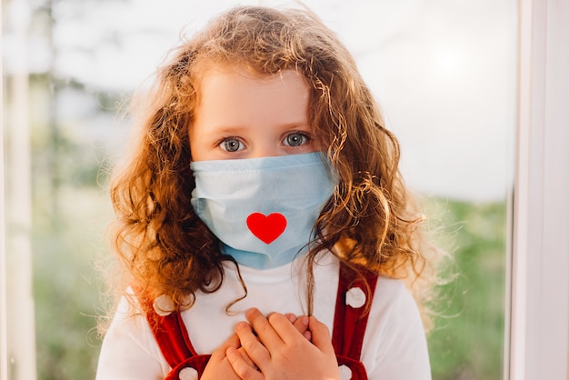 Close up portrait of little girl sitting on window sill wearing a health mask with a red heart on it as a way to show appreciation and to thank all essential employees during Covid-19 pandemic