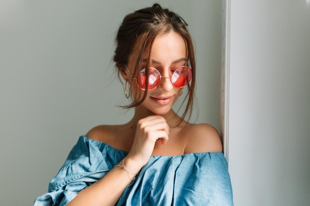 Close up portrait of lovable cute woman with collected dark\
hair wearing pink round glasses is trichina her chin and looking\
down in sunlight