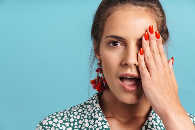 Close up portrait of a lovely excited young woman wearing shirt and bright earrings standing isolated over blue wall, looking 