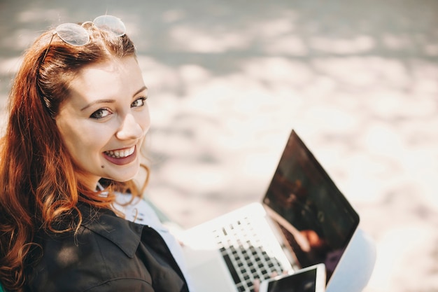 웃는 어깨 너머로 카메라를보고 공원에 앉아있는 동안 그녀의 노트북에서 일하는 빨간 머리를 가진 사랑스러운 젊은 더하기 크기 여자의 초상화를 닫습니다.