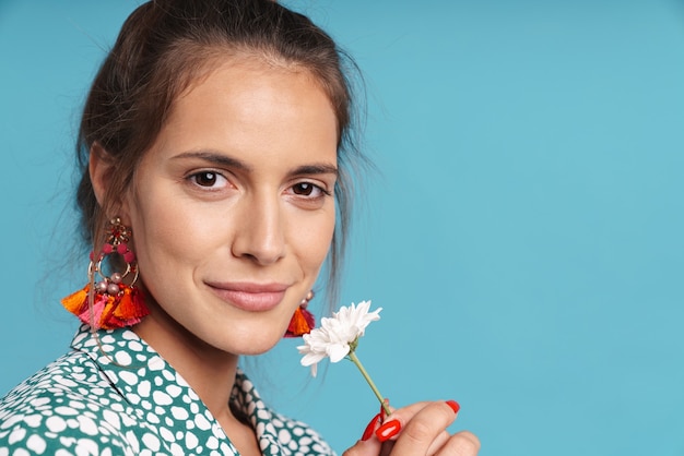 Close up portrait of a lovely young woman wearing shirt and bright earrings standing isolated over blue wall, holding chamomile