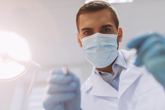 Close up portrait of male dentist in medical mask holding dental tools in modern dental clinic