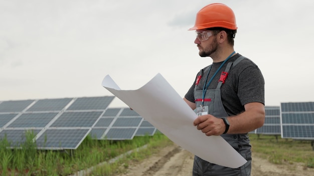 太陽光発電所の紙の計画の設計を学ぶ保護ヘルメットの男性の電気労働者の肖像画を閉じます。クリーンエネルギー。業界。グリーンエネルギーの概念。