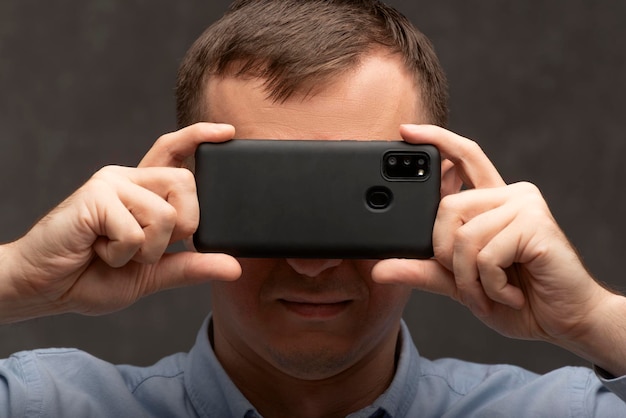 Close up portrait of man with phone near his face Young man on gray background holding mobile smartphone near eyes