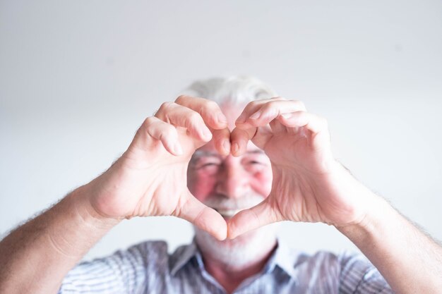 Photo close up and portrait of mature man and senior doing an heart with his fingers and hand in front of the camera - happy pensioner having fun enjoying - white background