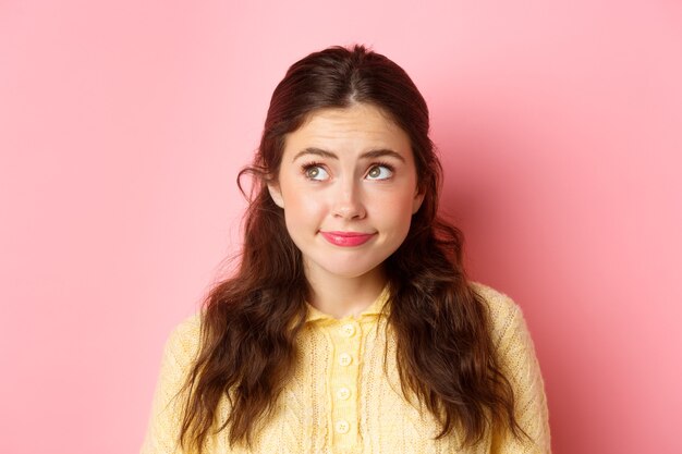 Close up portrait of modest cute girl, smiling and looking shy at upper left corner, standing thoughtful, wearing make up, pink wall.