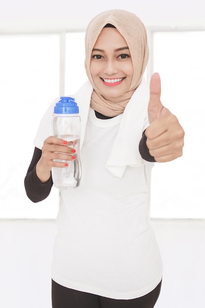 Close up portrait of muslim sporty woman holding a bottle of mineral water and giving thumbs up