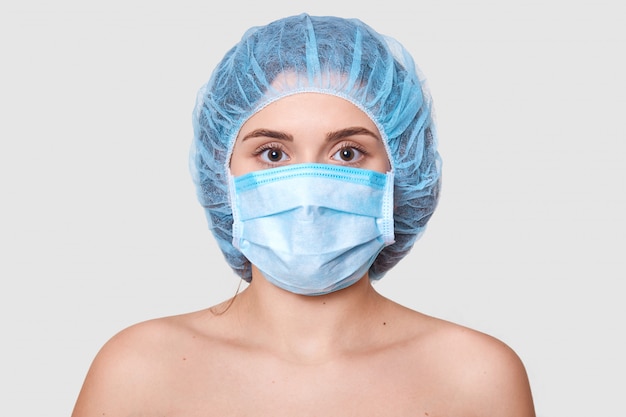 Close up portrait of naked woman standing isolated over white wall, wearing medical cap and mask, preparing for operation, looking seriously. Medicine concept.