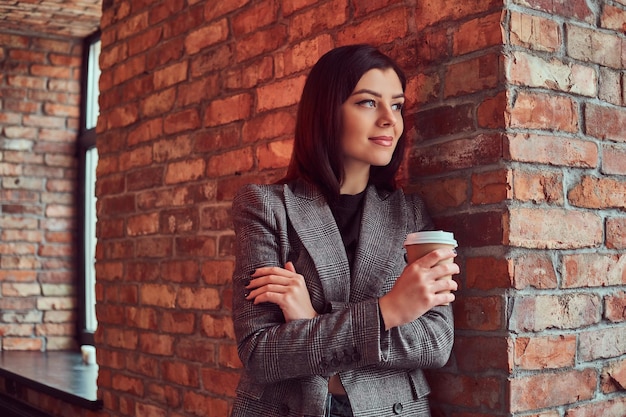 회색 우아한 재킷을 입은 관능적인 브루네트 여성의 클로즈업 초상화는 로프트 인테리어가 있는 방에서 벽돌 벽에 기대어 테이크아웃 커피 한 잔을 들고 있습니다.