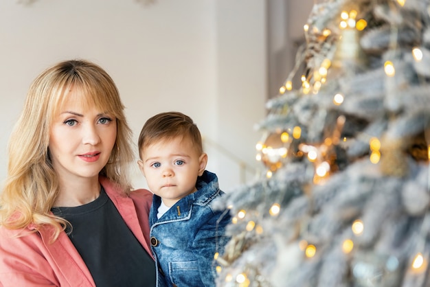 사진 크리스마스 트리 근처에 서 있는 세련된 어머니와 한 살짜리 아이의 클로즈업 초상화. 크리스마스 축하. 섣달 그믐 날. 육아 및 육아.