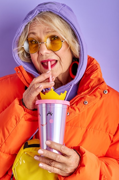 Фото Портрет крупным планом очаровательной пожилой женщины, пьющей сок с трубочкой, красивая женщина в ярких солнцезащитных очках под капотом смотрит в камеру