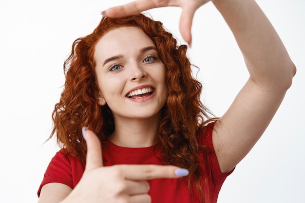 Крупным планом портрет красивой рыжей девушки с вьющимися волосами и естественным освещением, делает жест и улыбается, изображает что-то на белом