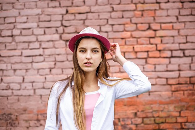 写真 背景としてピンクのレンガの壁の近くの帽子の美しいスタイリッシュな子供の女の子の肖像画をクローズアップ