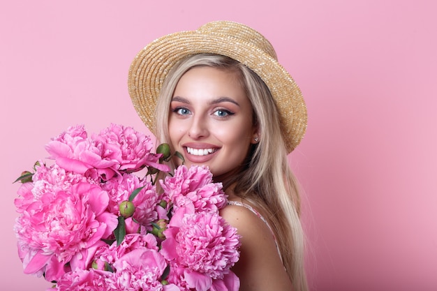 ピンクの上に牡丹の花束を保持している麦わら帽子の美しい若い女性のクローズアップの肖像画