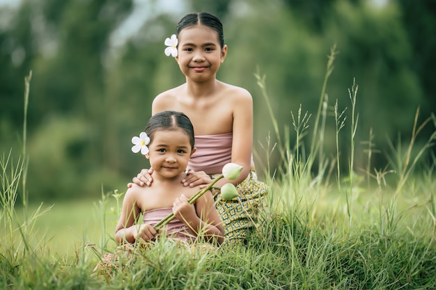 クローズアップ、タイの伝統的な衣装でかわいい妹と妹の肖像画と牧草地に座っている彼女の耳に白い花を置く、笑顔、兄弟愛の概念、コピースペース