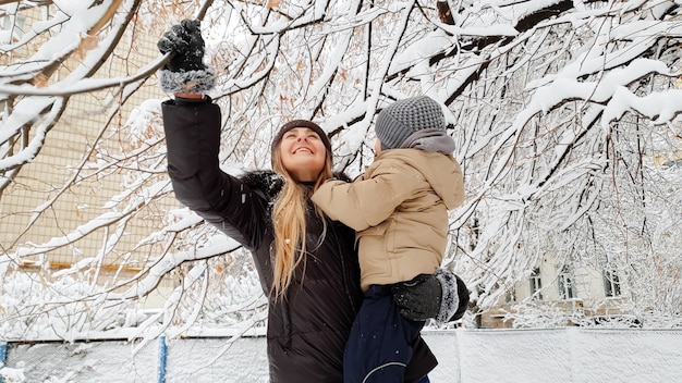 公園の遊び場で雪に覆われた木と遊ぶジャケットと帽子の彼女のかわいい息子と女性の肖像画をクローズアップ