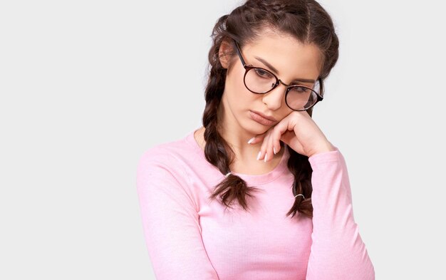 Close up portrait of overworked young brunette woman leans on hand has tired facial expression wears eyewear and pink casual blouse isolated over white background Shot of pretty sad female on gray