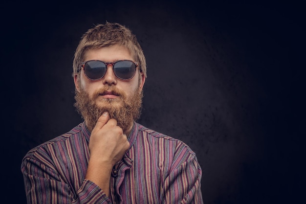 Close-up portrait of a pensive bearded redhead guy wearing sunglasses dressed in an old-fashioned shirt. Isolated on a dark background.