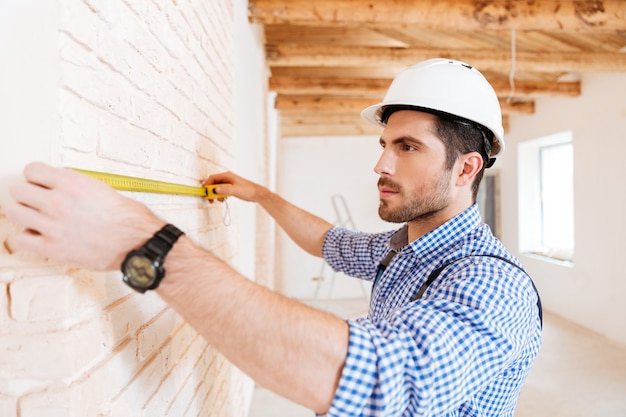 Close-up portrait of a pensive builder measuring wall using yellow type indoors