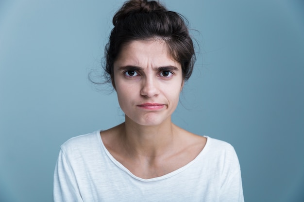 Close up portrait of a pensive unsatisfied pretty young brunette woman wearing white t-shirt standing isolated over blue background