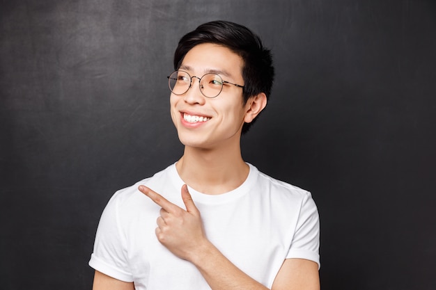 Il ritratto del primo piano del ragazzo asiatico sveglio vago soddisfatto in vetri individua qualcosa di fresco, indicando e guardando l'angolo in alto a sinistra con l'espressione soddisfatta e il sorriso raggiante,