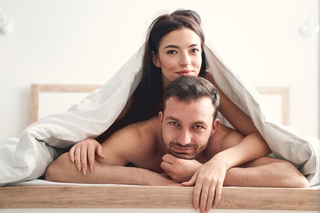Крупным планом портрет довольных молодоженов кавказской молодой пары под одеялом, глядя вперед
