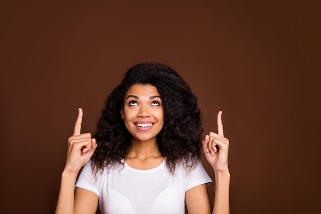 Close up portrait of positive funky afro american girl promoter point up index finger copyspace sect suggest ads promo wear white t-shirt.